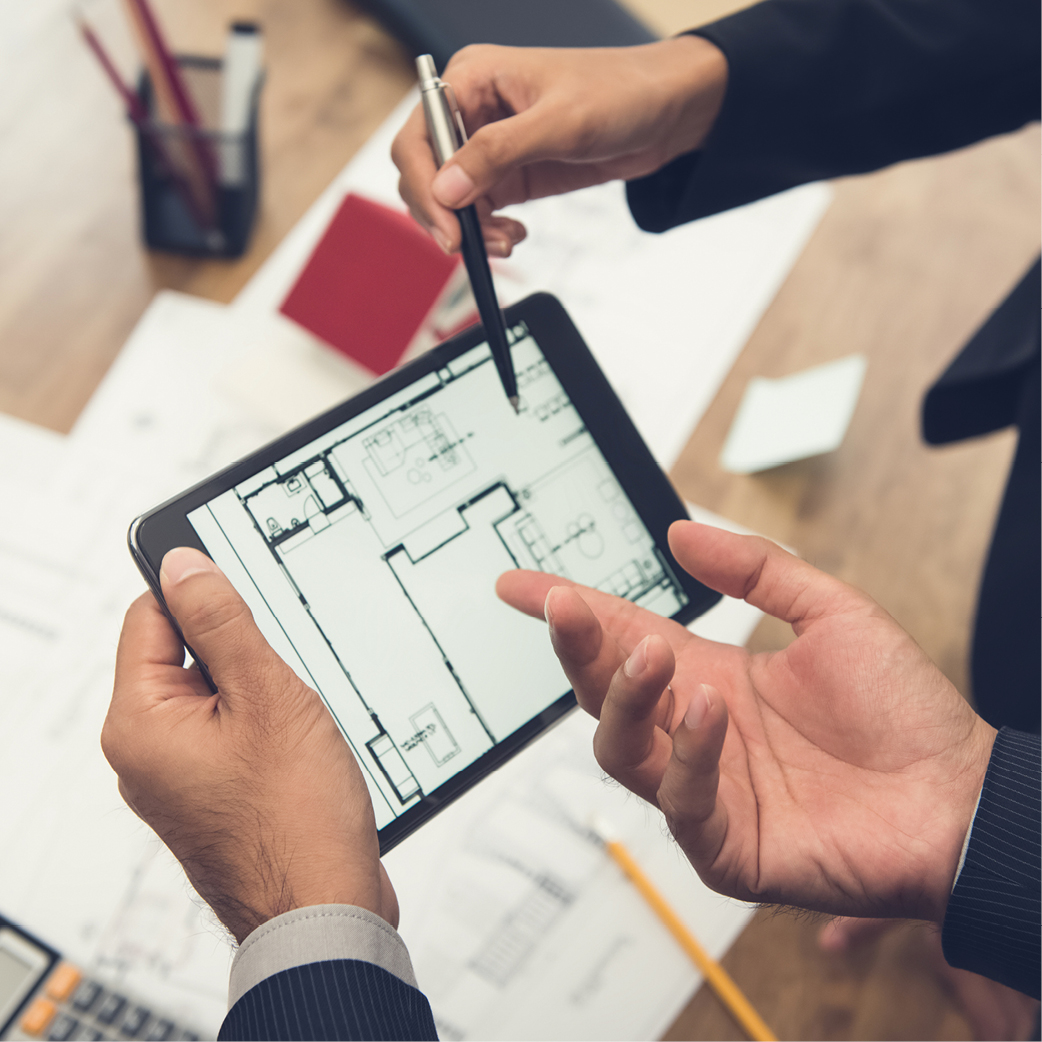 Zwei Hände halten ein Tablet, auf dem ein Grundriss abgebildet ist. Eine Hand zeigt mit einem Stift auf Details des Plans. Im Hintergrund sind Arbeitsmaterialien wie Blätter und Notizen sichtbar.