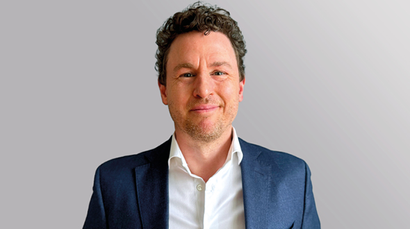 A man with curly hair smiles at the camera, wearing a dark blazer over a white shirt. The background is a soft gradient, emphasizing his friendly expression.