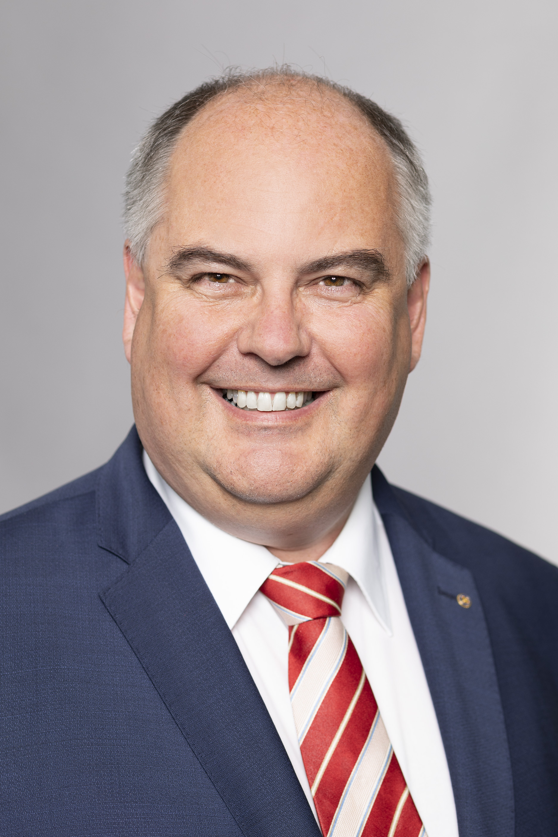 Porträt eines lächelnden Mannes mit kurzen grauen Haaren, der einen dunkelblauen Anzug und eine rot-gestreifte Krawatte trägt. Der Hintergrund ist grau. Der Mann strahlt Freundlichkeit und Selbstbewusstsein aus.