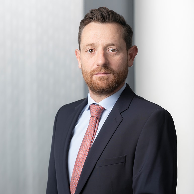 A man with a beard and short hair is dressed in a dark suit with a light blue shirt and a patterned tie. He is standing against a softly blurred, light background, looking directly at the camera with a serious expression.