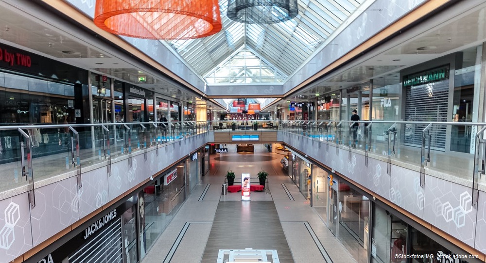 Eine moderne Einkaufsstraße in einem Einkaufszentrum, mit gläsernem Dach und mehreren Geschäften auf beiden Seiten. In der Mitte gibt es eine große offene Fläche, und die Wände sind mit einem hellen Design verziert. Einige Besucher sind im Hintergrund zu sehen.