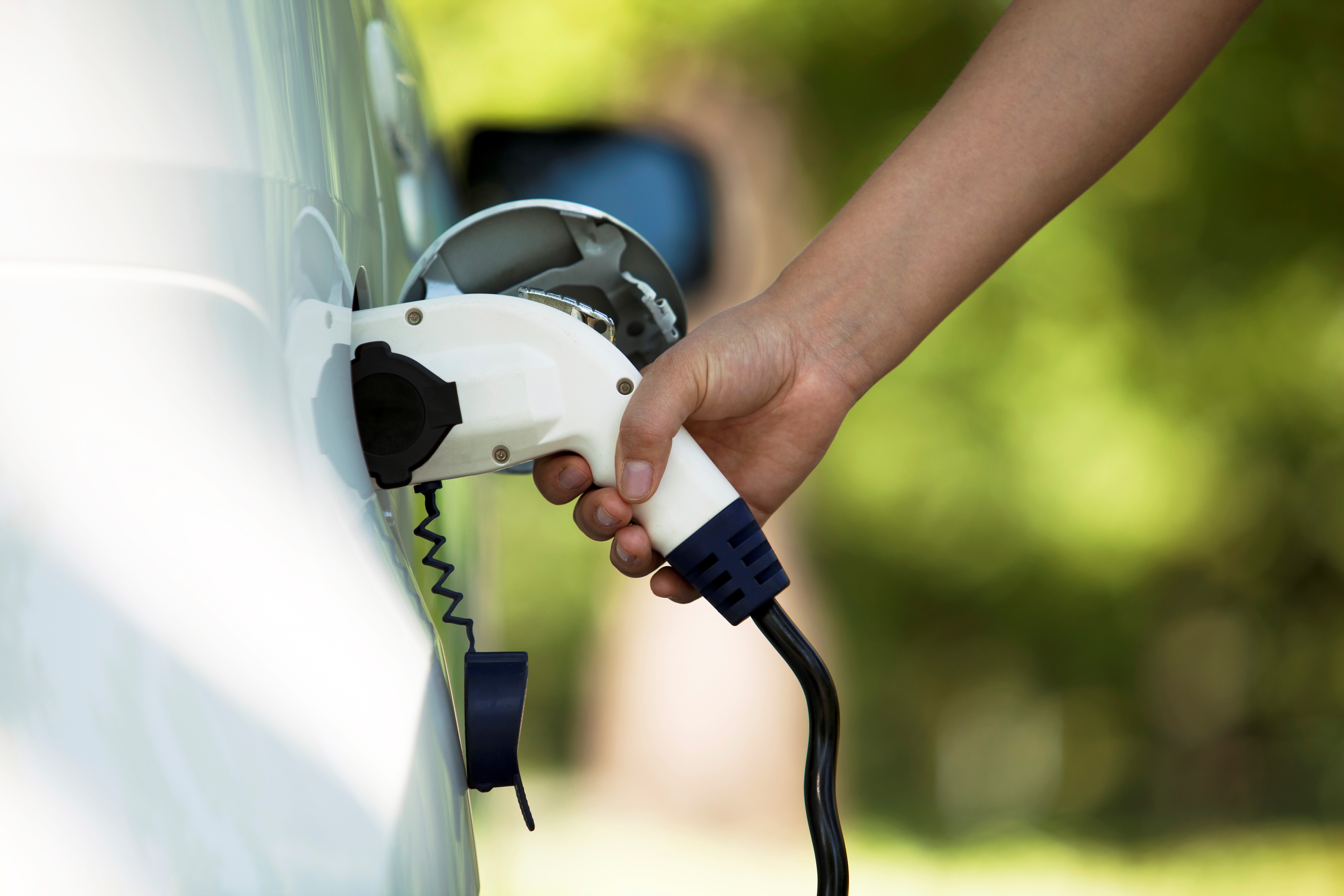 A hand is holding an electric vehicle charging plug, preparing to connect it to a charging port on the car. The background features blurred greenery, indicating an outdoor setting.