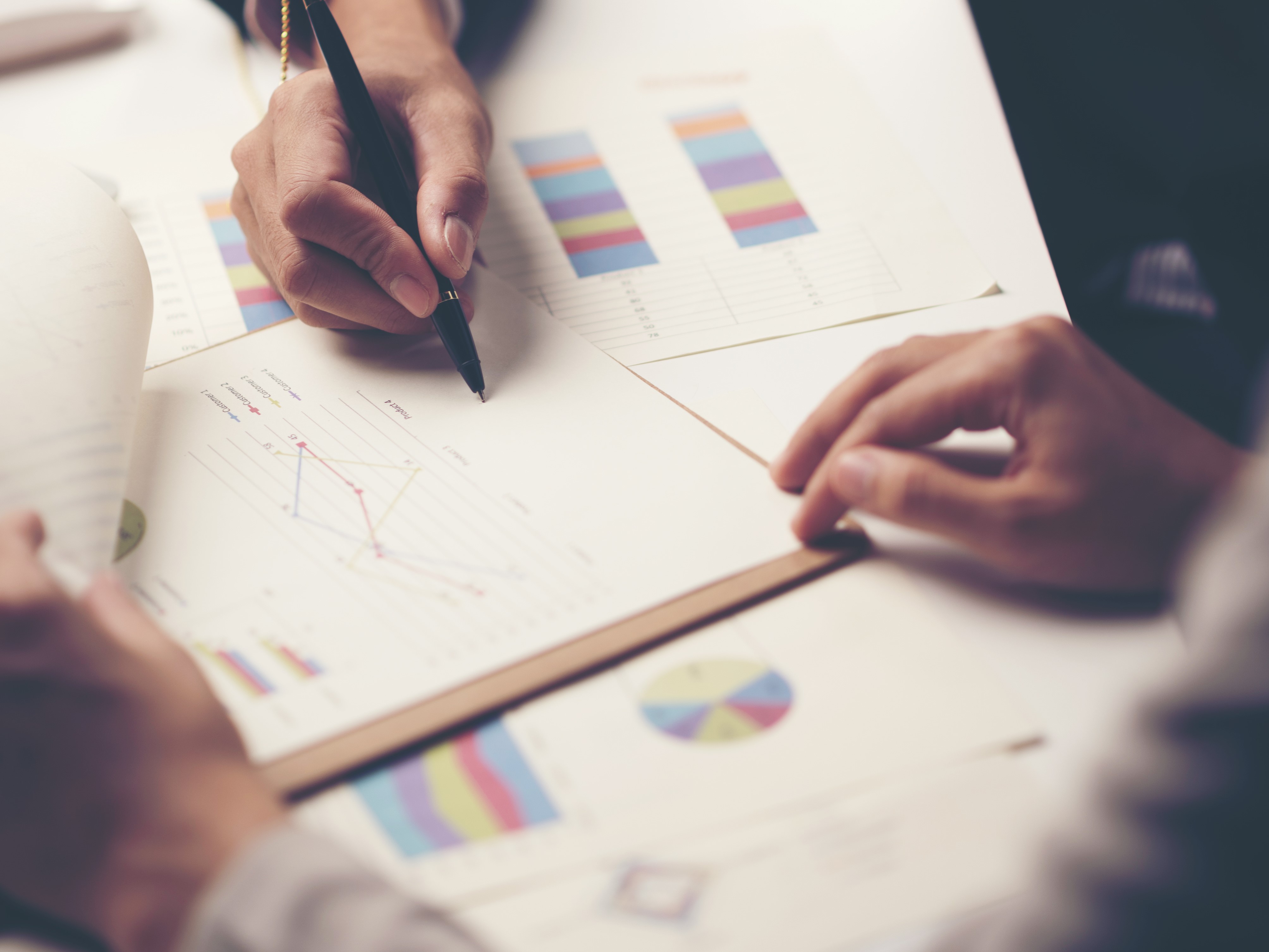 Zwei Hände befinden sich auf einer Arbeitsfläche, umgeben von Dokumenten mit Diagrammen und Grafiken. Eine Hand hält einen Stift und macht Notizen auf einem Blatt mit einem Liniendiagramm. Visuelle Datenanalysen scheinen diskutiert zu werden.