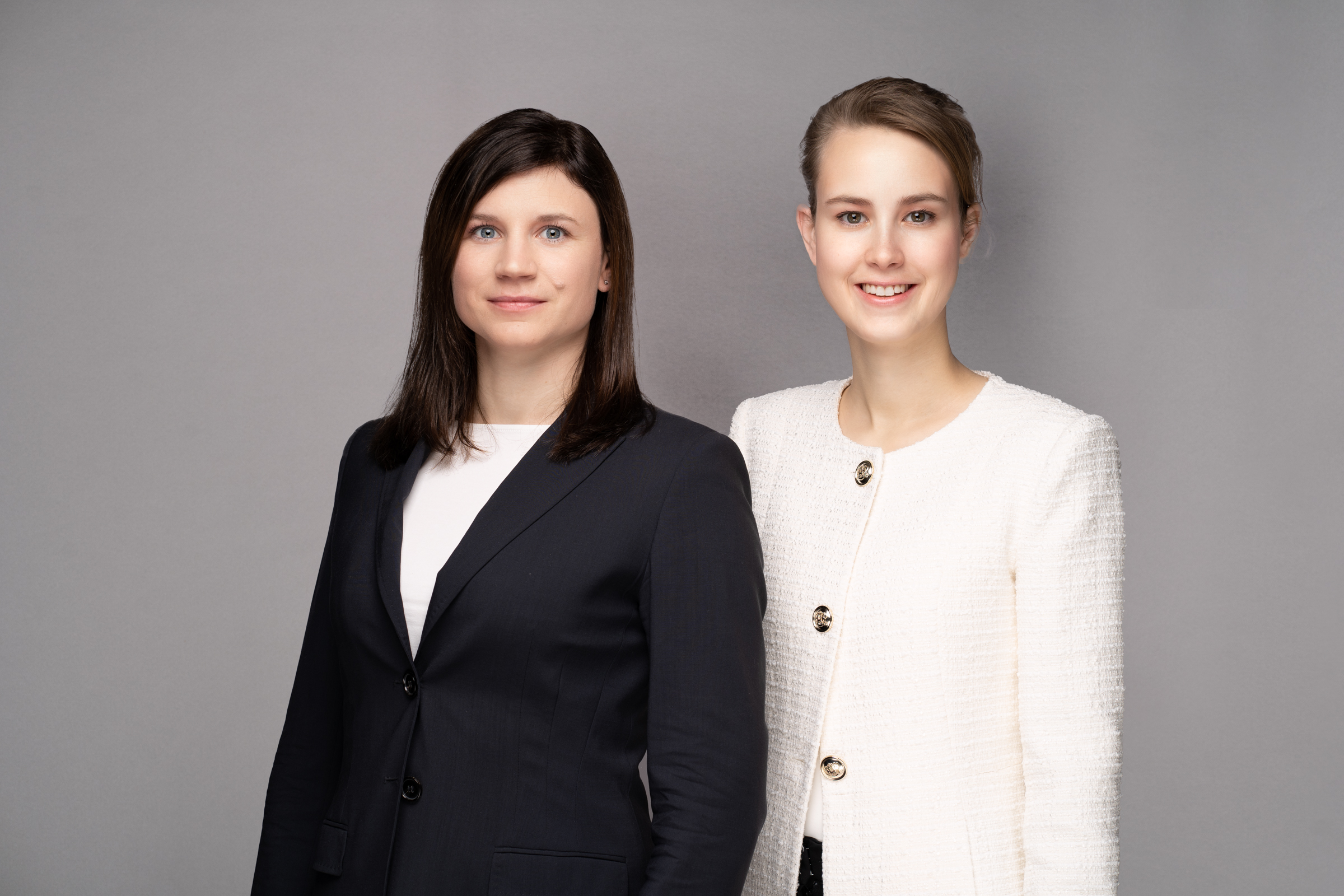 Two women stand side by side, both wearing formal attire. The woman on the left has long dark hair and is dressed in a dark blazer over a white top. The woman on the right has light hair styled back and is wearing a light-colored, textured jacket with buttons.