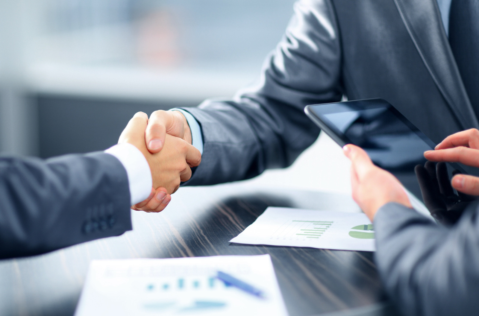 Two business professionals are shaking hands across a table, symbolizing an agreement or partnership. One person holds a tablet, while documents with graphs are laid out on the table. The setting suggests a modern office environment focused on collaboration.