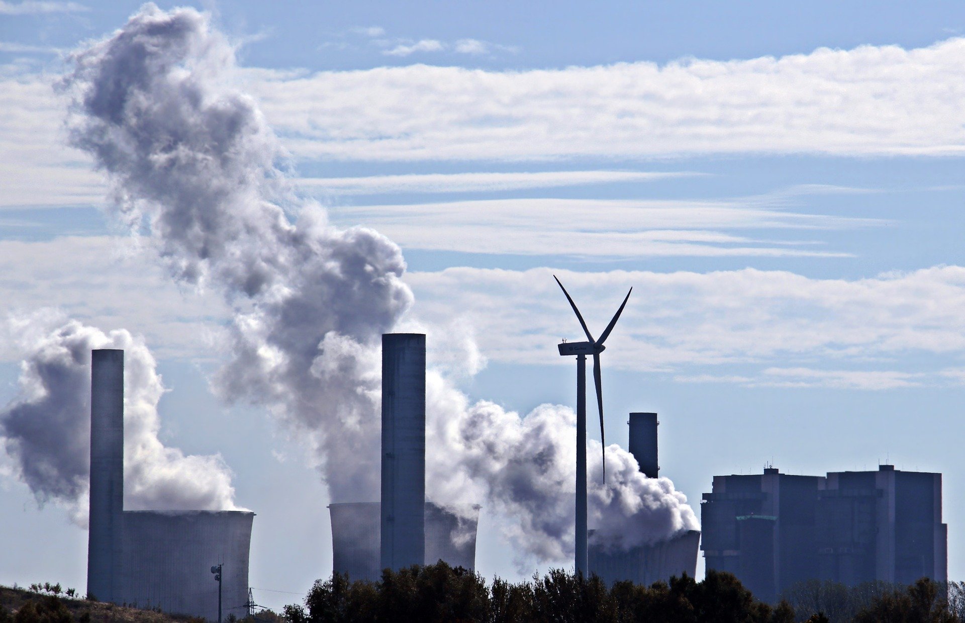Das Bild zeigt eine industrielle Landschaft mit mehreren Kühl- und Schornsteintürmen, aus denen Rauch aufsteigt. Im Vordergrund steht ein Windrad, das erneuerbare Energie symbolisiert. Die Szenerie kontrastiert zwischen umweltschädlicher und sauberer Energieerzeugung.
