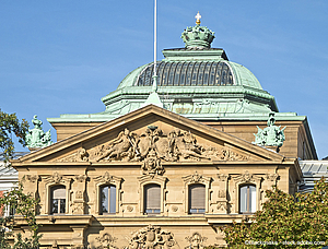 Das Bild zeigt die Fassade eines historischen Gebäudes mit einer kunstvoll gestalteten Front. Besonders hervorstechend sind der kupferne Kuppeldach und die reich verzierten Reliefs, die Figuren und Ornamentik zeigen. Der Himmel ist klar und blau.