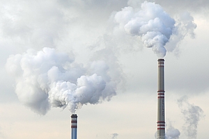 Image features two tall smokestacks emitting large plumes of white smoke against a cloudy sky, indicating industrial activity. The smoke suggests air pollution concerns associated with manufacturing processes.
