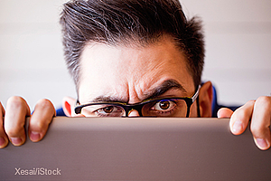 Ein Mann mit Brille schaut gespannt über den Rand eines Laptops. Seine Stirn ist leicht gerunzelt, und seine Augen zeigen Neugier oder Besorgnis. Der Hintergrund ist unscharf, was den Fokus auf den Mann und sein Gesicht verstärkt.