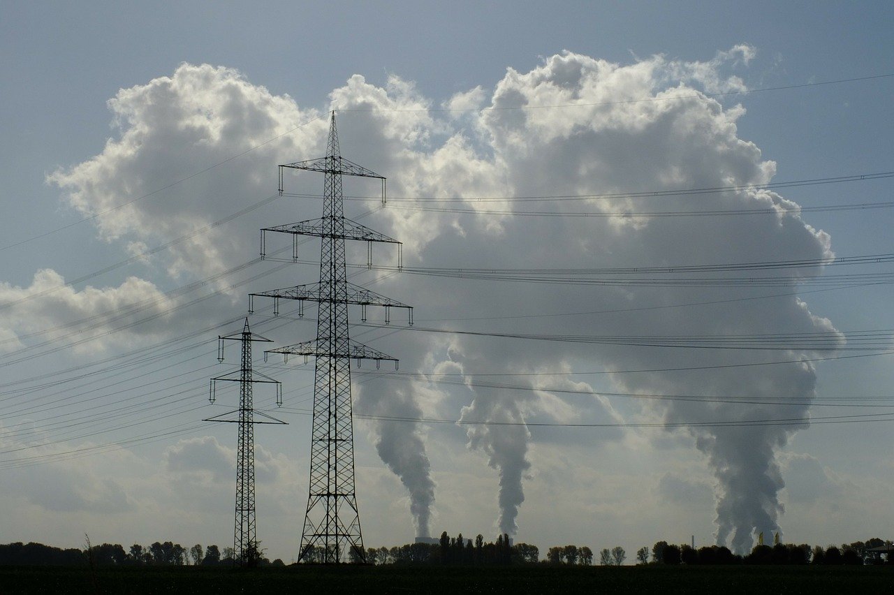 Ein starker Strommast steht im Vordergrund, umgeben von weiteren Masten. Im Hintergrund sind hohe Rauchschwaden zu sehen, die aus einem Industriegebiet aufsteigen. Der Himmel ist bewölkt und vermittelt eine düstere Atmosphäre.