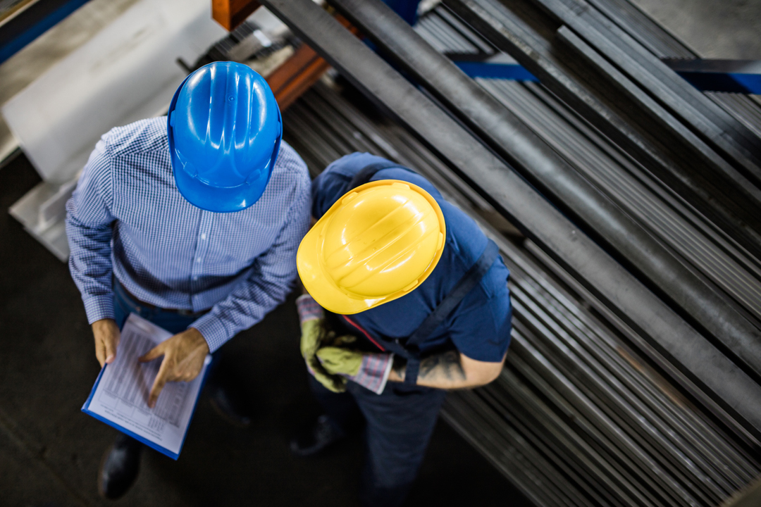 Zwei Arbeiter in einem industriellen Umfeld sind zu sehen, einer trägt einen blauen und der andere einen gelben Schutzhelm. Sie stehen über einer Liste von Dokumenten und scheinen über etwas zu diskutieren, während metallene Stangen im Hintergrund angeordnet sind.