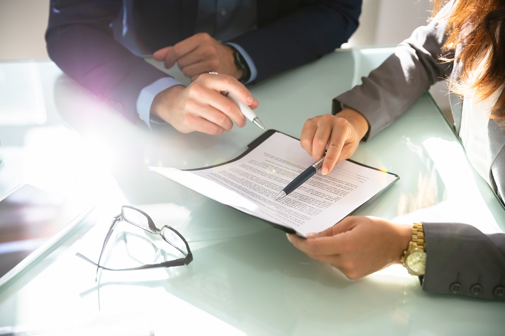 Zwei Personen sitzen an einem Tisch und betrachten ein Dokument. Eine Hand hält einen Kugelschreiber, während die andere auf den Text deutet. Die Szene impliziert eine Besprechung oder Vertragsunterzeichnung. Brillenzubehör liegt auf dem Tisch.