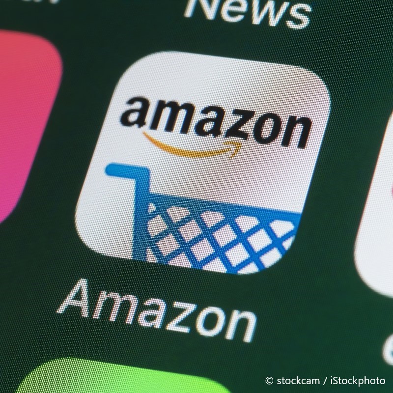 Das Bild zeigt das Amazon-App-Symbol auf einem Bildschirm. Es besteht aus dem Wort "Amazon" in schwarzer Schrift über einem Einkaufswagensymbol mit einer blauen Linie. Der Hintergrund ist hell und der Einkaufswagen ist in einem stilisierten, grafischen Design gehalten.
