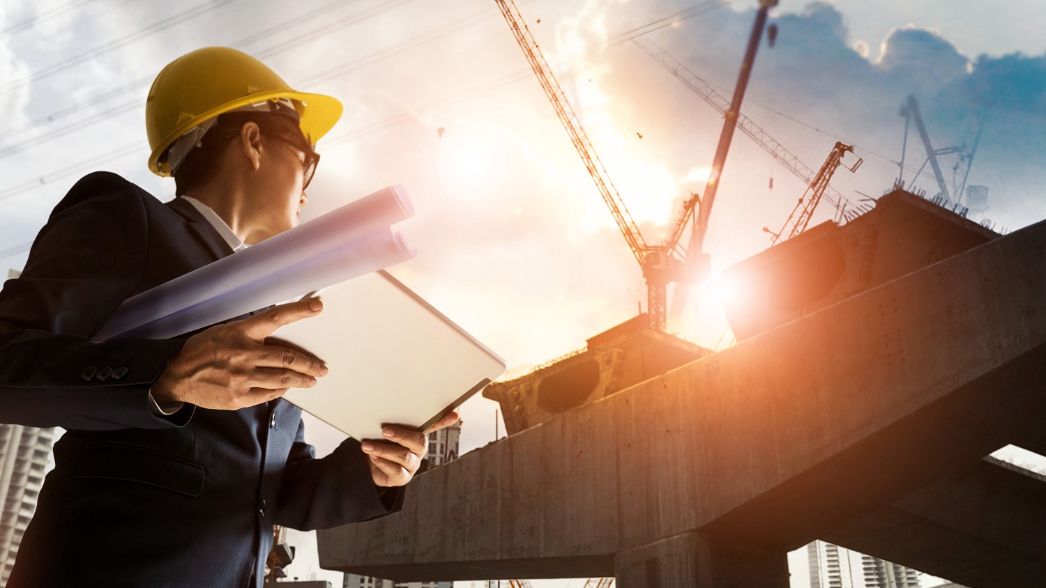 A man in a suit and hard hat stands on a construction site, holding blueprints and a tablet. He gazes at a large structure being built, with cranes and scaffolding visible against a cloudy sky. The scene conveys a sense of progress in construction and planning.