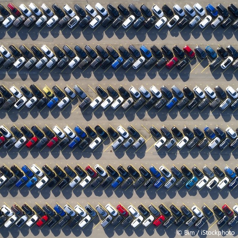 Eine Luftaufnahme eines Parkplatzes, auf dem Fahrzeuge in mehreren Reihen und Spalten parken. Die Autos sind in verschiedenen Farben angeordnet, darunter schwarz, weiß, blau und rot. Die Anordnung der Autos ist ordentlich und gleichmäßig, was einen strukturierten Überblick über den Parkplatz ermöglicht.