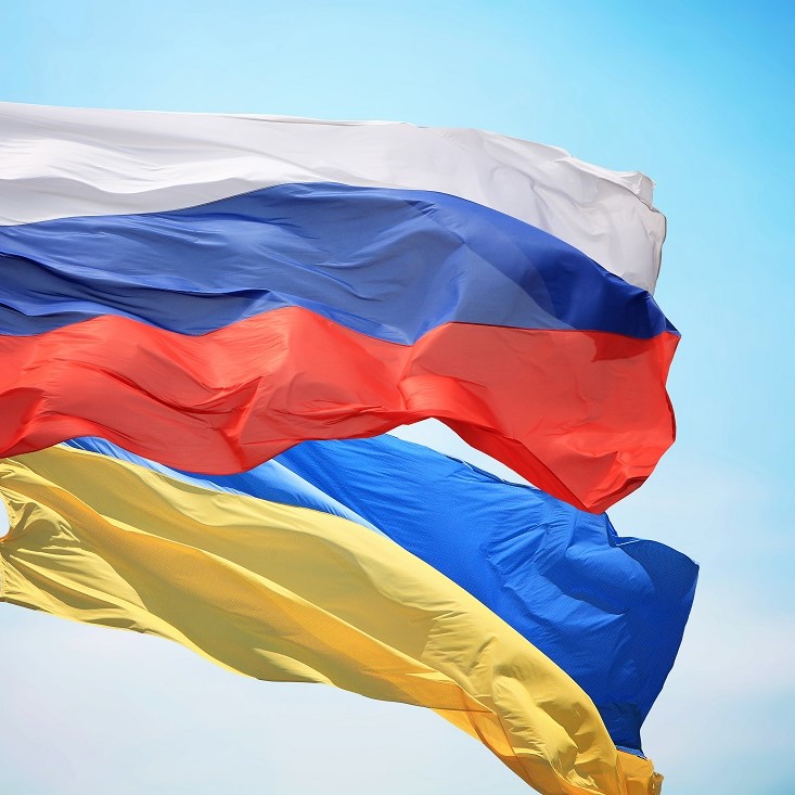 Drei Fahnen wehen im Wind: Die oberste ist weiß, die mittlere zeigt rot, blau und weiß (Russland) und die unterste ist blau und gelb (Ukraine). Der Hintergrund ist ein klarer blauer Himmel.