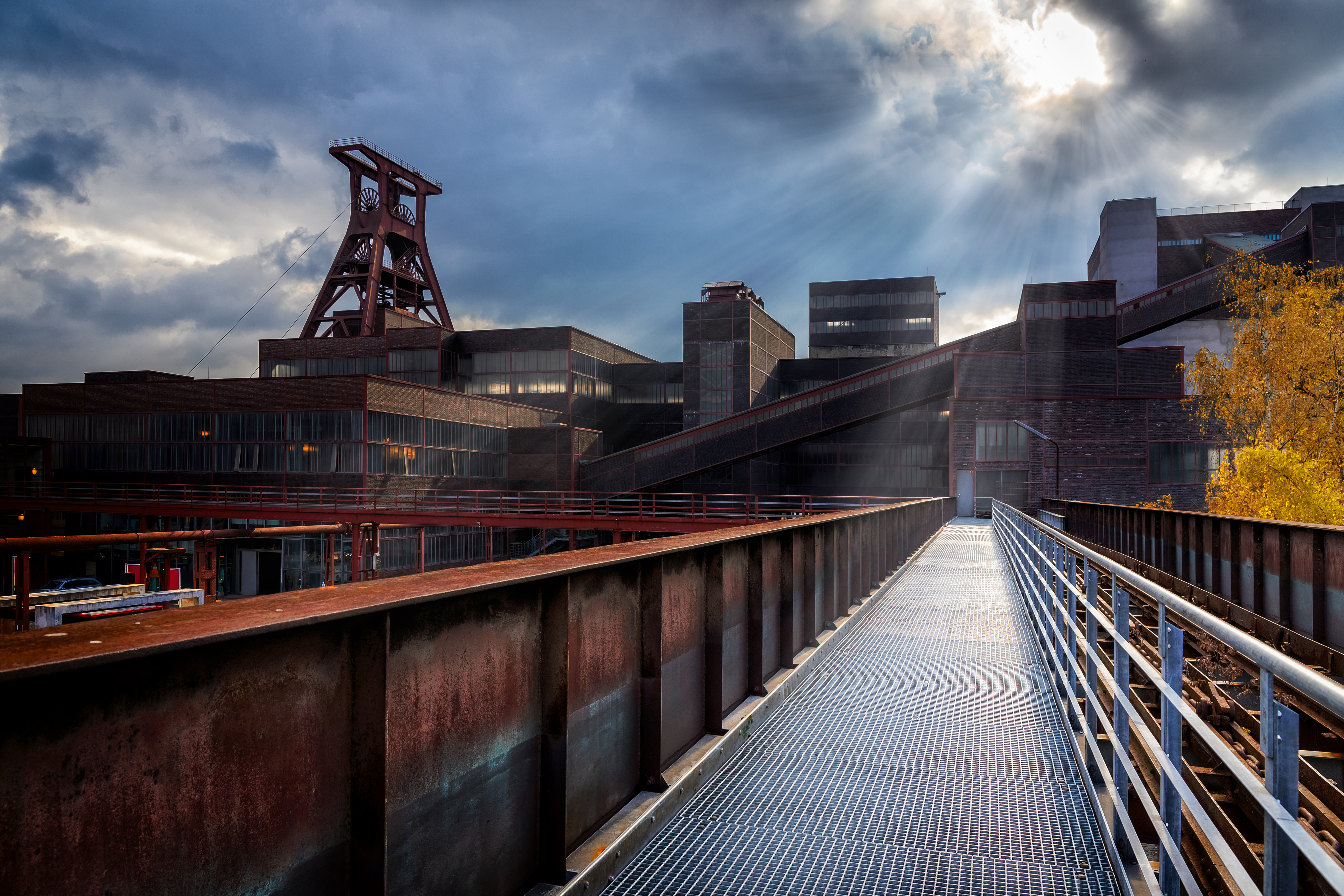 Eine alte Industrieanlage mit einem markanten Förderturm und modernen Gebäuden, unter einem bewölkten Himmel mit Lichtstrahlen. Im Vordergrund steht eine Metallbrücke, die zur Anlage führt, umgeben von Pflanzen und einer urbanen Landschaft.