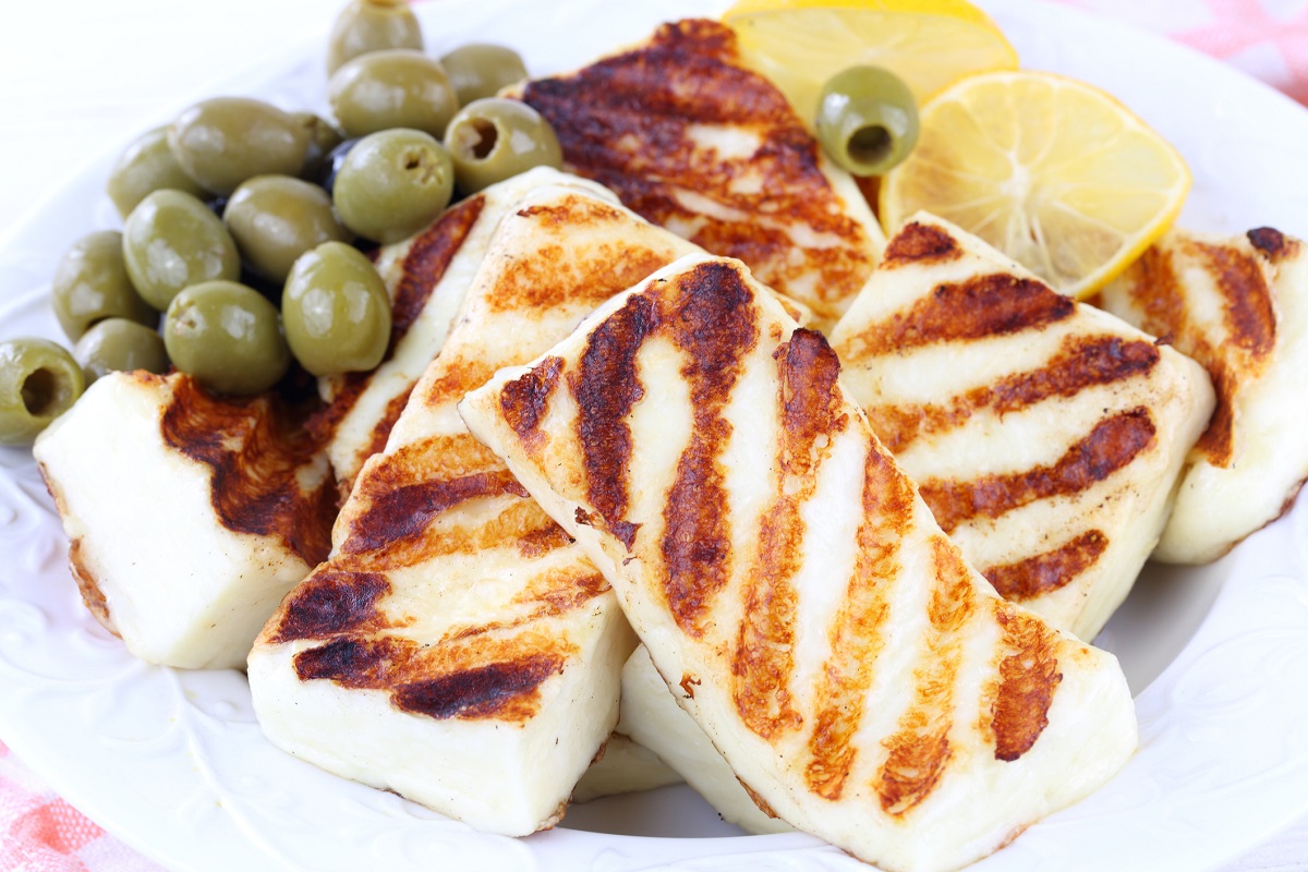 Gegrillter Halloumi-Käse liegt auf einem weißen Teller, umgeben von grünen Oliven und frischen Zitronenscheiben. Der Käse hat eine goldbraune, gestreifte Oberfläche und wirkt appetitlich.
