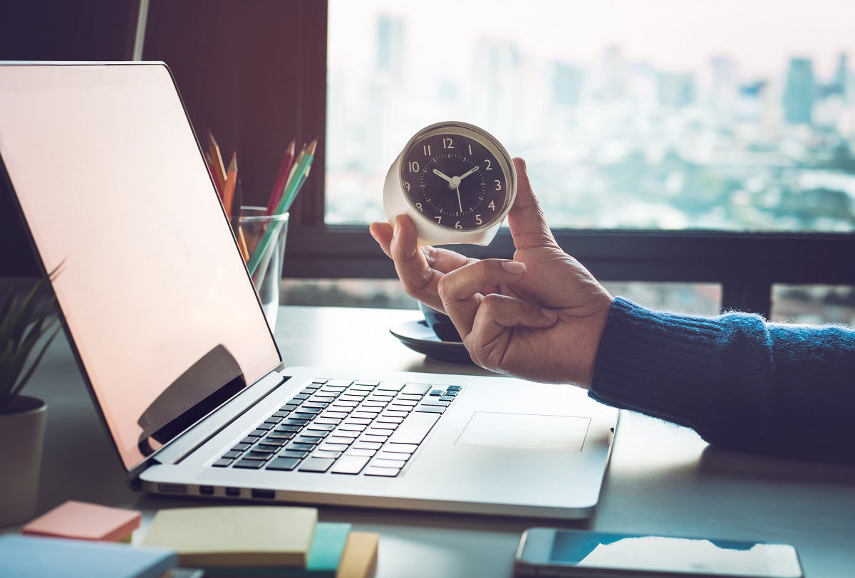 Eine Hand hält eine Uhr in der Nähe eines offenen Laptops. Der Hintergrund zeigt große Fenster mit Stadtansicht. Der Schreibtisch ist mit bunten Notizen und einem Becher mit Stiften ausgestattet. Die Szene vermittelt eine arbeitsame Atmosphäre.