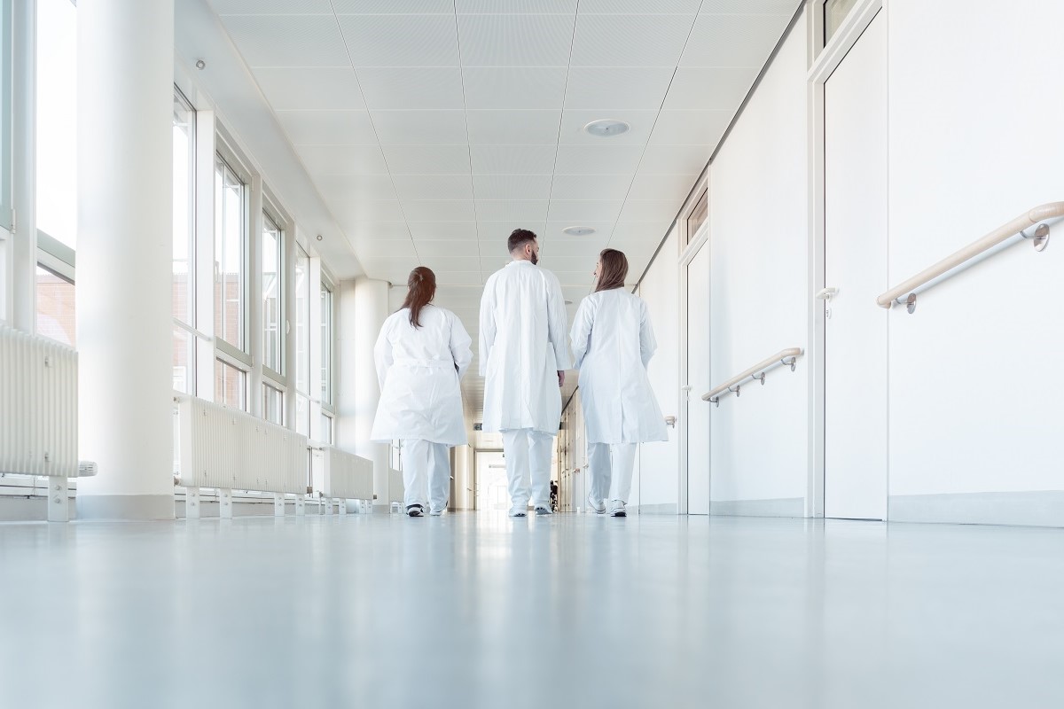 Three medical professionals in white lab coats walk together down a well-lit hospital corridor. The scene conveys a sense of teamwork and professionalism in a healthcare setting, with large windows allowing natural light to fill the space.