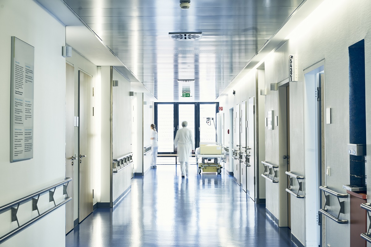 Ein heller Krankenhausflur mit mehreren Türen zu Patientenzimmern. Am Ende des Flurs sind große Fenster, die Tageslicht hereinlassen. Eine Person in einem weißen Kittel geht in Richtung Fenster, während eine weitere Person ebenfalls im Flur sichtbar ist.