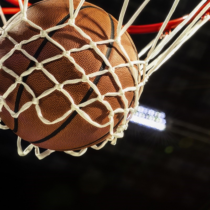 Ein Basketball schwebt in einem Basketballkorb, dessen Netz aus weißen Schnüren besteht. Die orangefarbene Oberfläche des Balls ist gut sichtbar. Im Hintergrund sind unscharfe Lichtquellen zu erkennen.