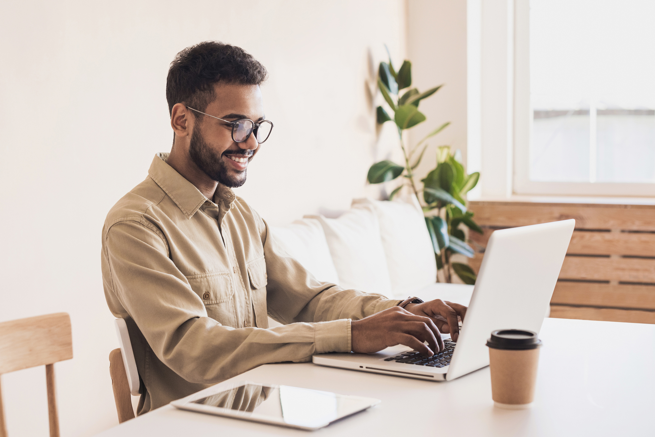 Ein junger Mann mit Bart und Brille sitzt an einem Tisch und arbeitet konzentriert an einem Laptop. Er trägt ein beige Hemd und lächelt. Neben dem Laptop steht ein Kaffeebecher. Im Hintergrund ist eine helle, freundliche Umgebung mit Pflanzen und einem Sofa zu sehen.
