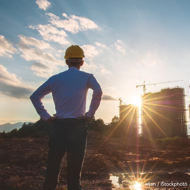 Eine Person mit einem Schutzhelm steht mit verschränkten Händen auf einer Baustelle und schaut auf ein großes Bauprojekt im Sonnenlicht. Die Sonne scheint hinter den Gebäuden und erzeugt eine strahlende Atmosphäre mit Wolken am Himmel.