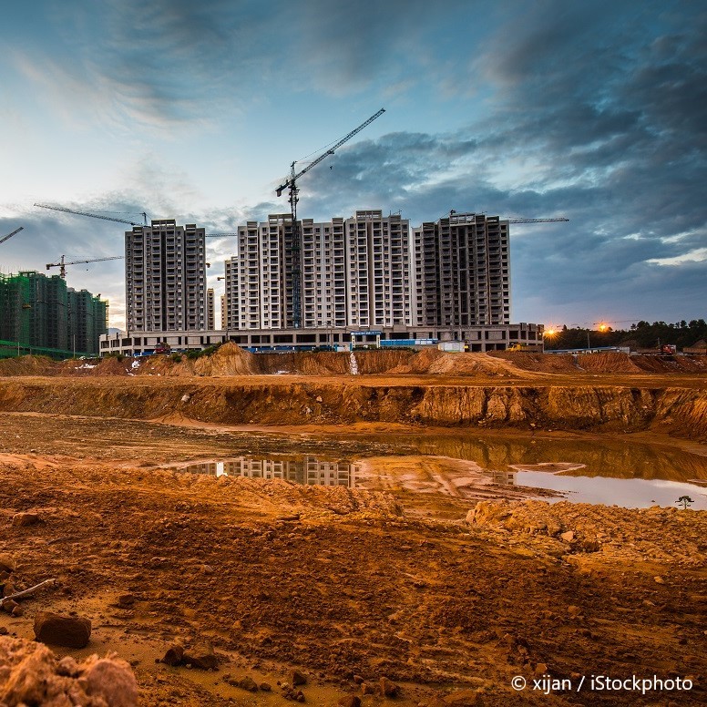 Eine Baustelle mit mehreren hohen Wohngebäuden im Hintergrund, die teilweise errichtet sind. Im Vordergrund sind unebene Erdmassen und eine Wasseransammlung sichtbar. Kräne sind über den Gebäuden positioniert, und der Himmel zeigt eine Mischung aus Wolken und Licht.