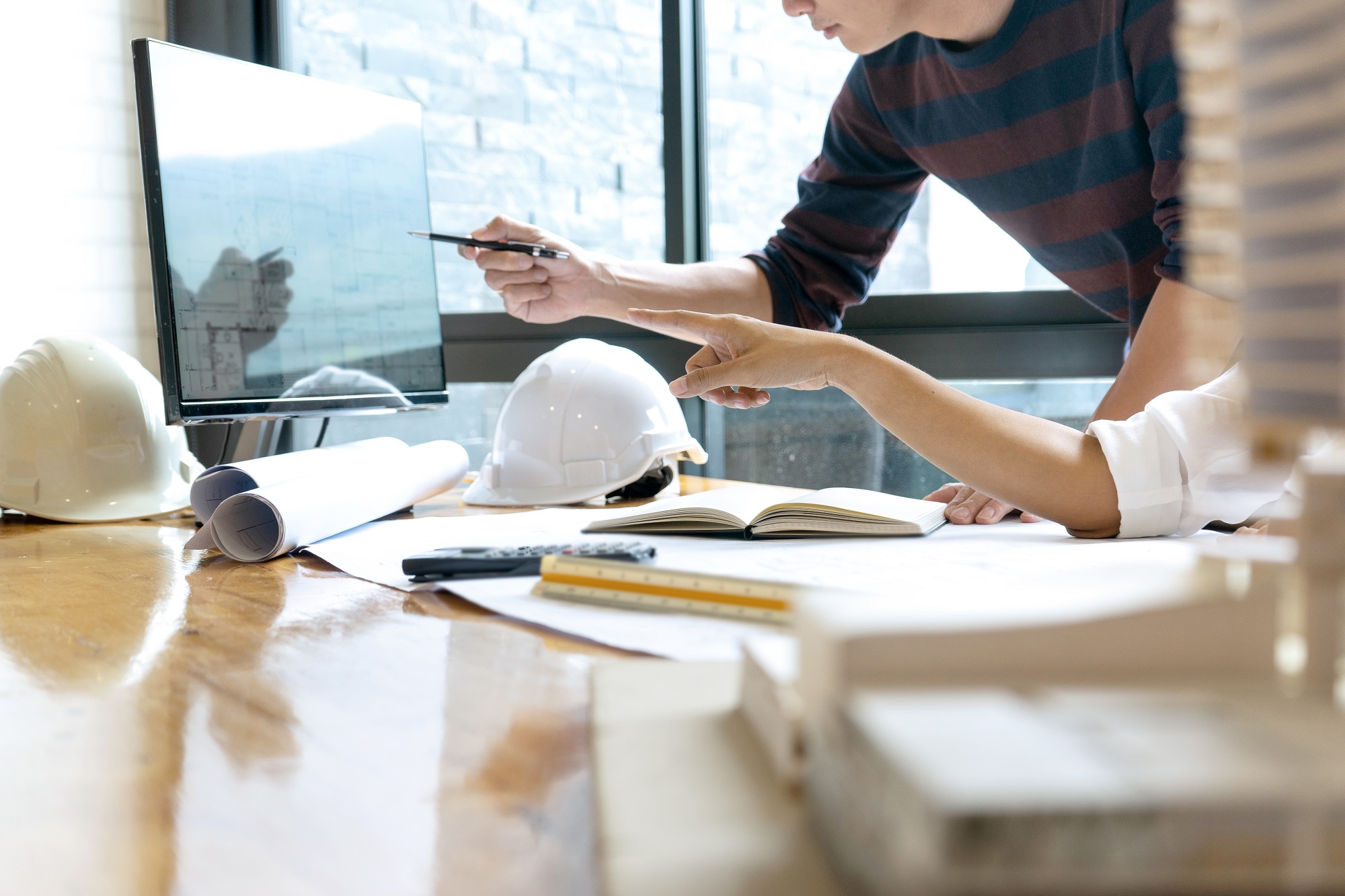 Two individuals are discussing plans in an office setting. One person is pointing at a computer screen displaying diagrams, while the other is holding a pen and gesturing. There are construction helmets, rolled blueprints, a notepad, and a calculator on the table.