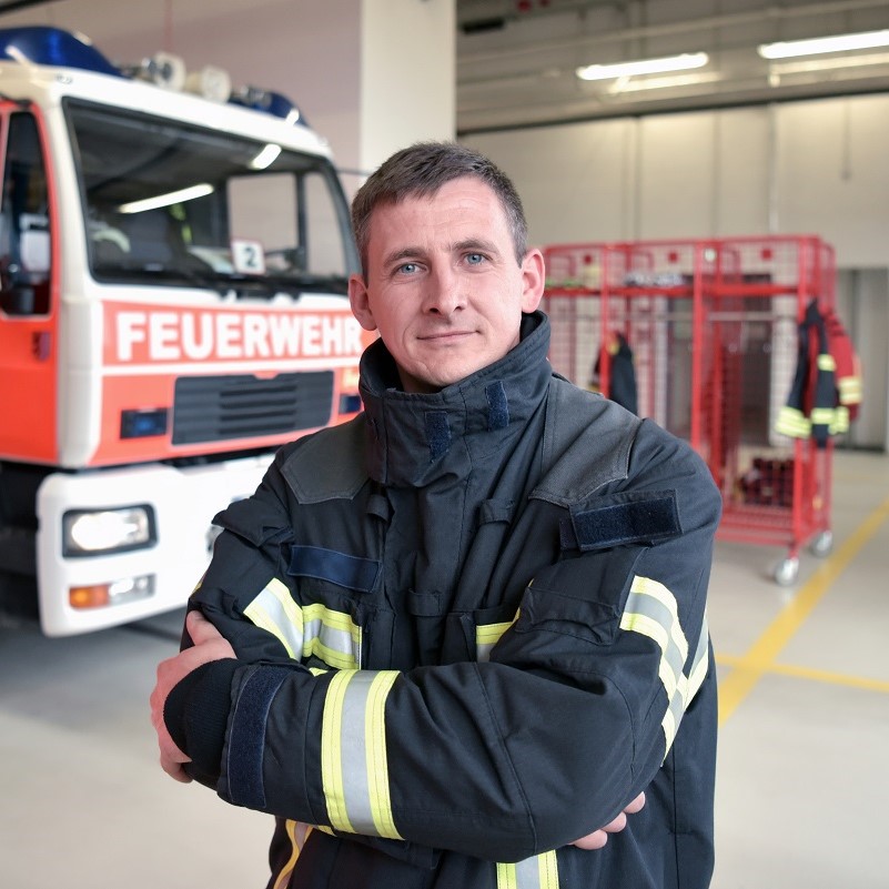 Ein Feuerwehrmann in Uniform steht mit verschränkten Armen vor einem Feuerwehrfahrzeug. Der Hintergrund zeigt weitere Feuerwehrausrüstungen in einem modernen Feuerwehrhaus. Die Atmosphäre vermittelt Professionalität und Bereitschaft zum Einsatz.