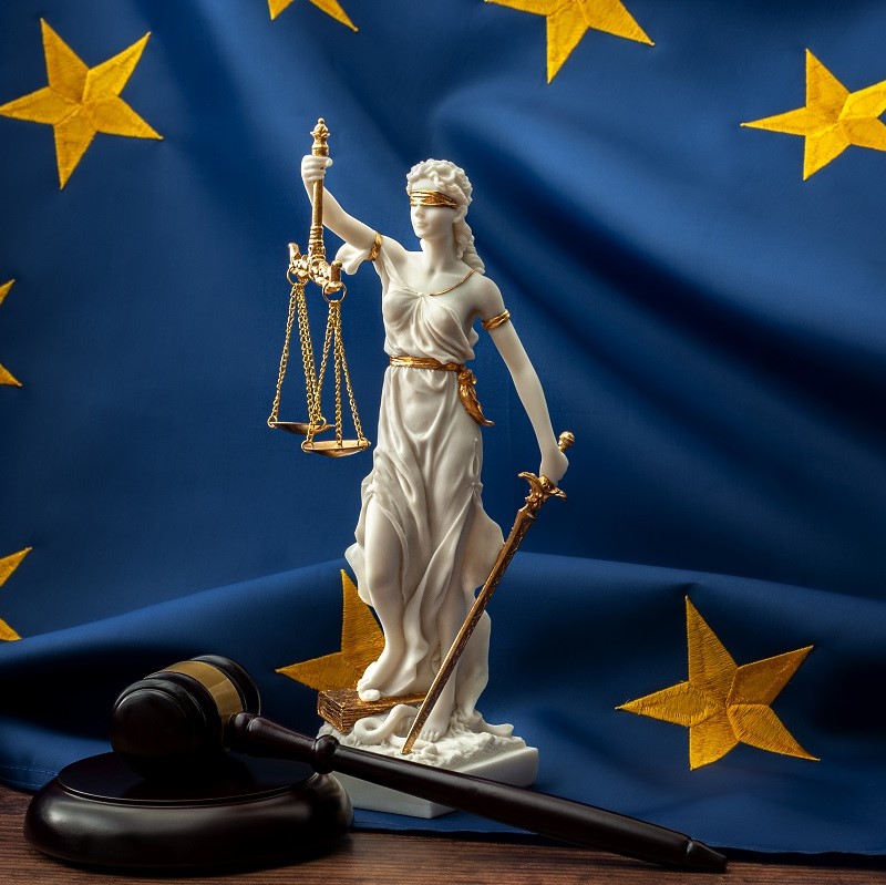 Eine Statue der Justitia mit verbundenen Augen hält Waage und Schwert. Im Hintergrund entfaltet sich eine europäische Flagge. Daneben liegt ein Richterhammer auf einem Tisch. Die Darstellung symbolisiert Gerechtigkeit und das Rechtssystem in Europa.