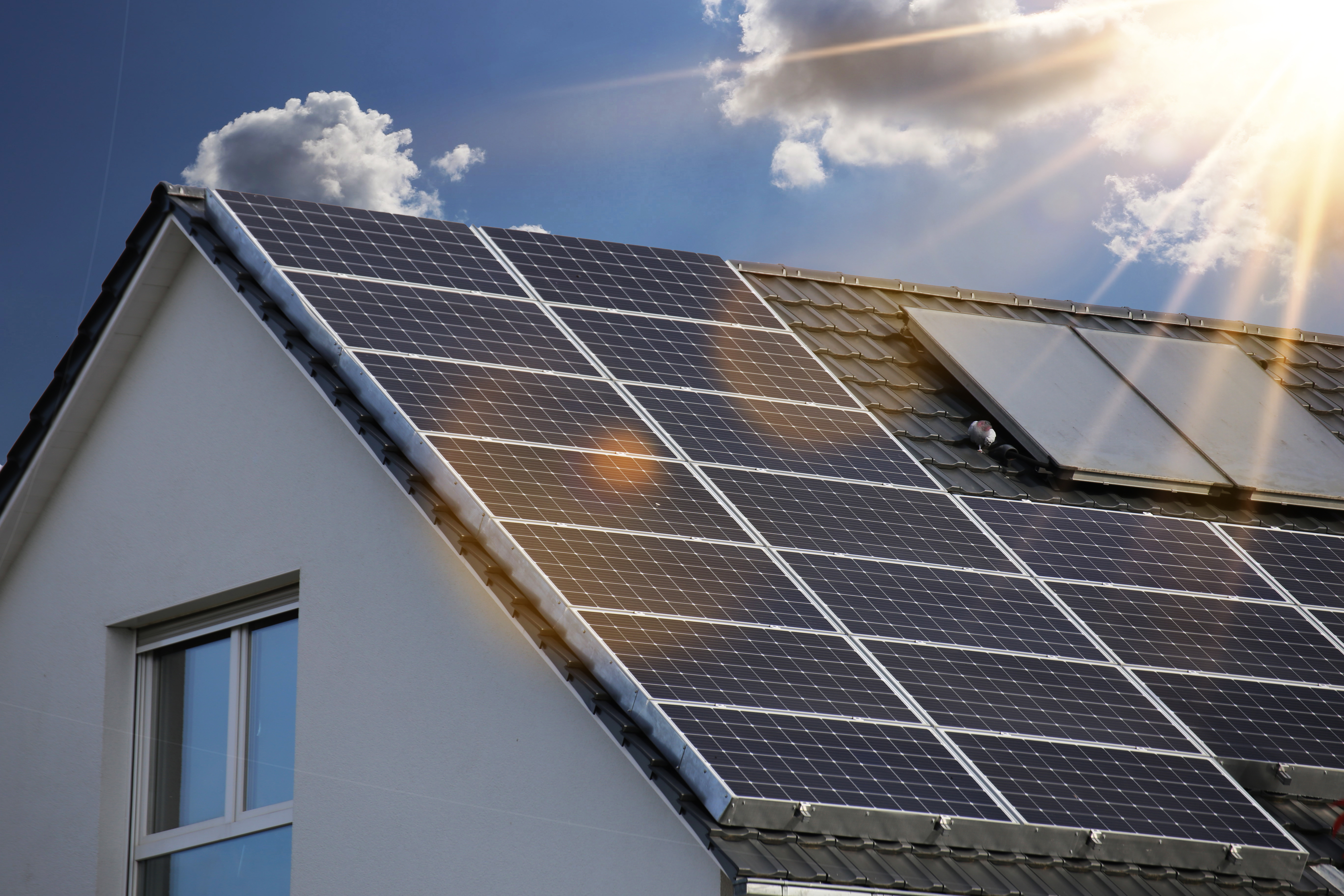 Ein Dach mit Solarpanelen ist unter einem blauen Himmel mit einigen Wolken zu sehen. Die Solarpanele sind gut sichtbar und reflektieren das Sonnenlicht, was auf die Nutzung von Solarenergie hinweist.