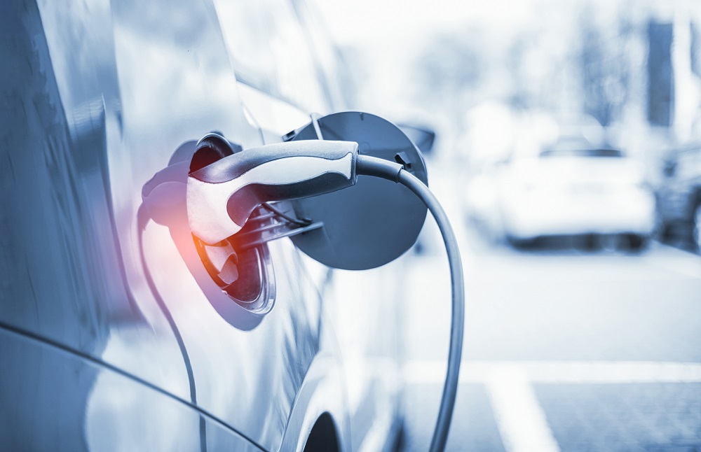 Ein Elektrofahrzeug ist an einer Ladestation angeschlossen. Der Ladestecker ist in die Ladebuchse des Autos gesteckt, während ein Kabel von der Ladestation zum Fahrzeug führt. Die Szene vermittelt das Thema Elektromobilität und umweltfreundliches Fahren.