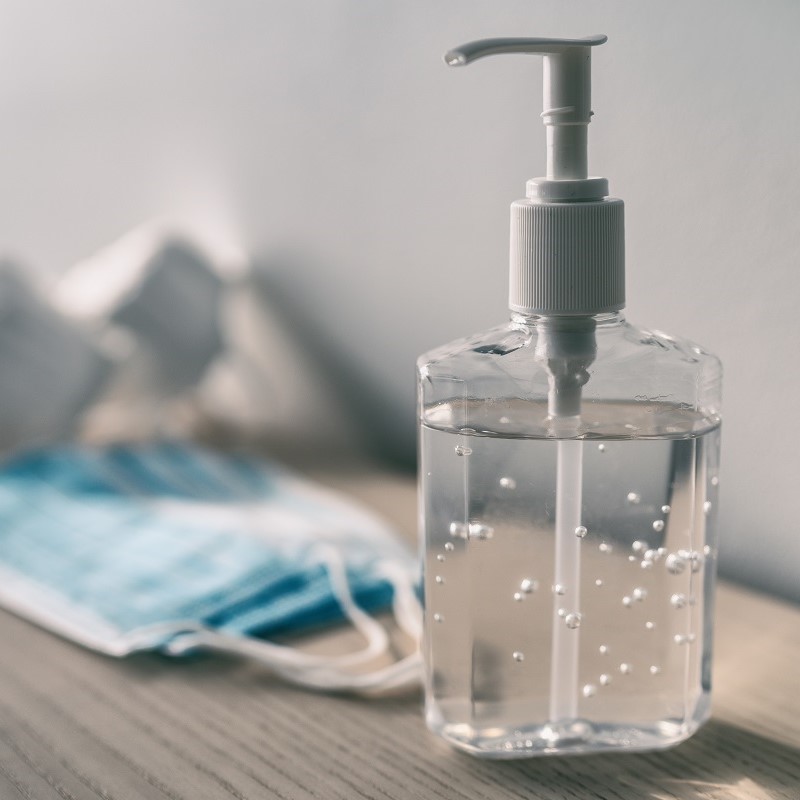 Eine Flasche mit Desinfektionsmittel steht auf einem Tisch. Die Flasche hat eine Pumpvorrichtung und enthält eine klare Flüssigkeit mit kleinen Bläschen. Im Hintergrund liegt eine blaue Maske.