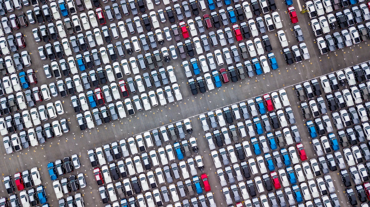 Eine Luftaufnahme eines Parkplatzes, der mit zahlreichen Autos gefüllt ist. Die Fahrzeuge sind hauptsächlich in verschiedenen Grautönen, mit einigen blauen und roten Autos, die sich in der Masse abheben. Die Parkplätze sind ordentlich angeordnet, sichtbar durch gelbe Markierungen auf dem Boden.