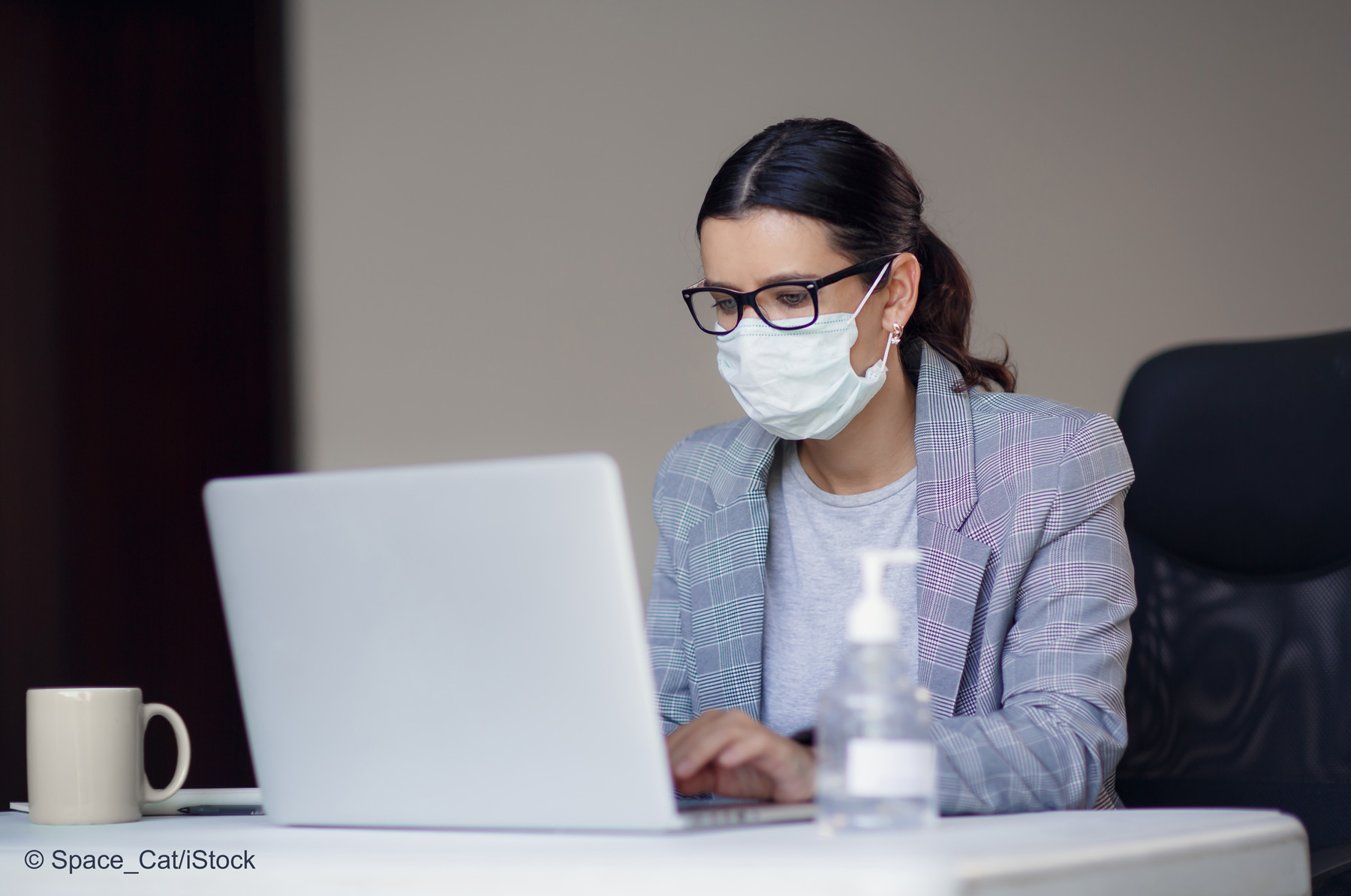 Eine Frau mit Brille und Mundschutz sitzt an einem Schreibtisch und arbeitet an einem Laptop. Neben ihr steht eine Flasche mit Handdesinfektionsmittel und eine Tasse. Der Hintergrund ist unscharf, um den Fokus auf die Frau und ihre Tätigkeit zu legen.