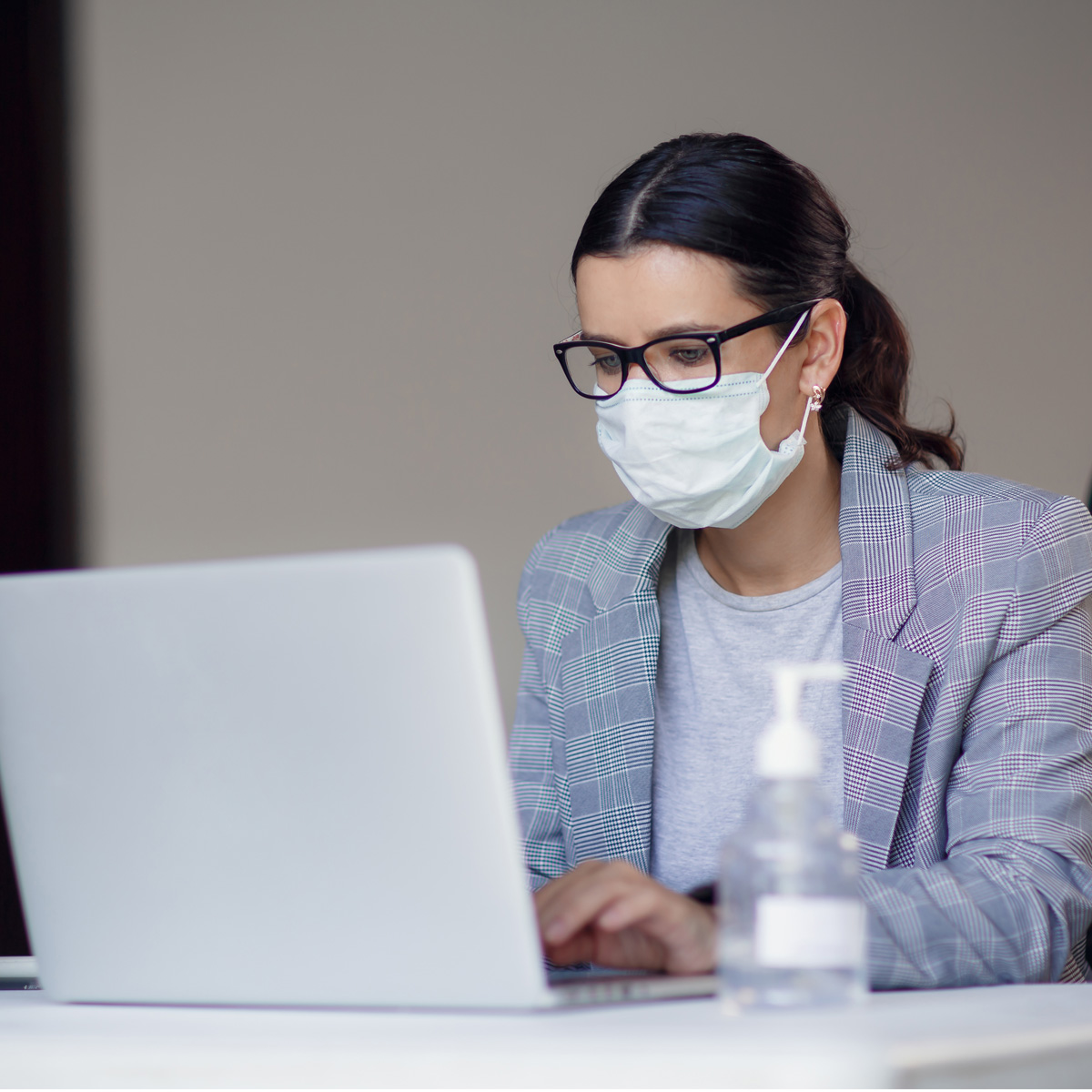 Eine Frau mit Brille trägt eine Mundmaske und arbeitet an einem Laptop. Sie sitzt an einem Tisch, auf dem eine Flasche Desinfektionsmittel steht. Der Hintergrund ist unscharf und neutral gehalten.