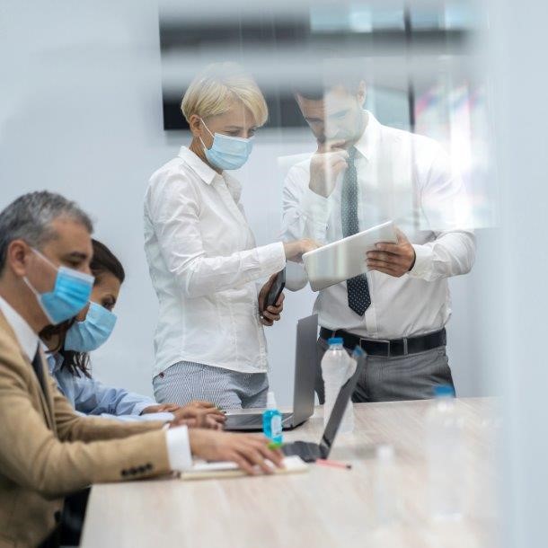 Eine Gruppe von fünf Personen in einem Konferenzraum, die Masken tragen. Zwei Personen stehen nebeneinander und schauen auf ein Tablet, während die anderen am Tisch arbeiten. Alle scheinen an einer Besprechung oder Präsentation beteiligt zu sein.