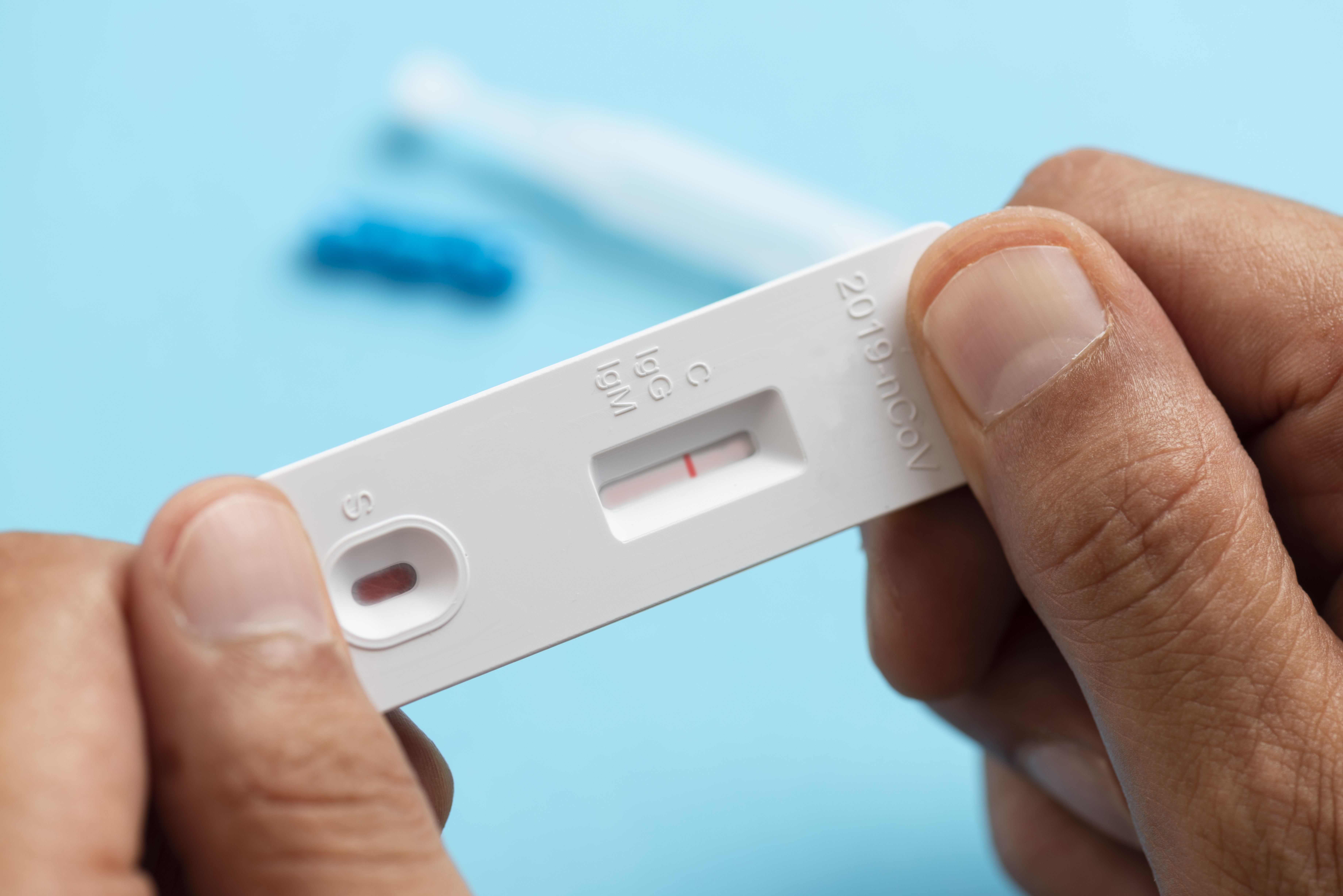 Eine Person hält einen COVID-19-Teststreifen, auf dem ein rotes Strichmuster sichtbar ist. Der Test wird vor einem blauen Hintergrund präsentiert. Der Streifen zeigt die Bezeichnungen C für Kontrolle und T für Test.