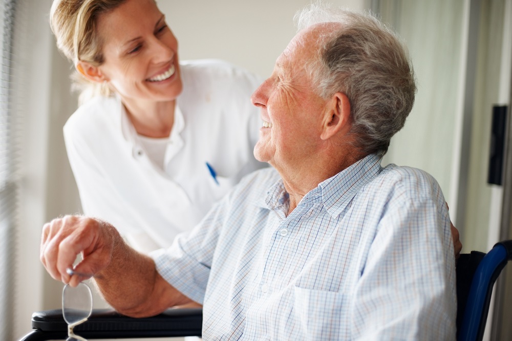 A smiling caregiver leans toward an elderly man seated in a wheelchair, creating a warm and friendly atmosphere. The caregiver appears engaged and attentive, while the man smiles back, suggesting a moment of connection and care in their interaction.