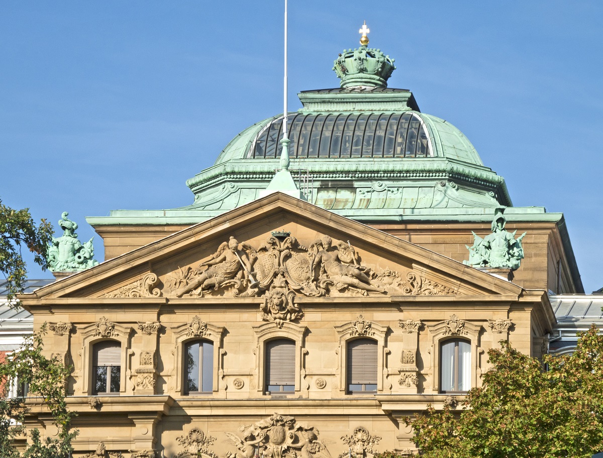 Das Bild zeigt die Fassade eines historisch gestalteten Gebäudes mit aufwendigen Reliefs und Fenstern. Ein gewölbtes Dach mit einer Kuppel in Kupferfarbe ist im Hintergrund sichtbar. Die Architektur vermittelt einen imposanten, klassischen Stil.