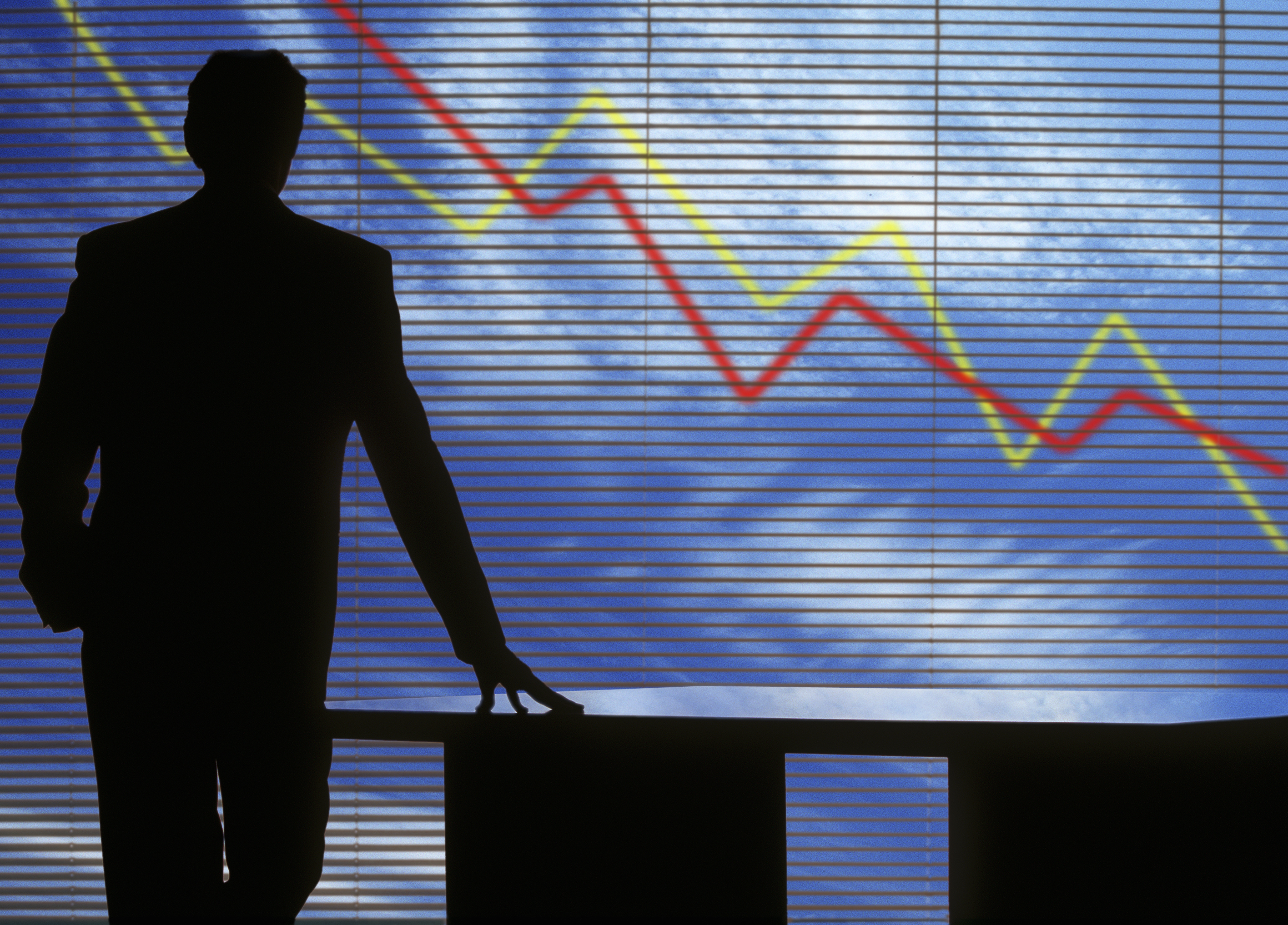 A silhouette of a person in a suit stands by a table, gazing at a blue background with a graph showing red and yellow lines, indicating fluctuating trends. The scene suggests a business or financial context. The background has horizontal blinds casting shadows.