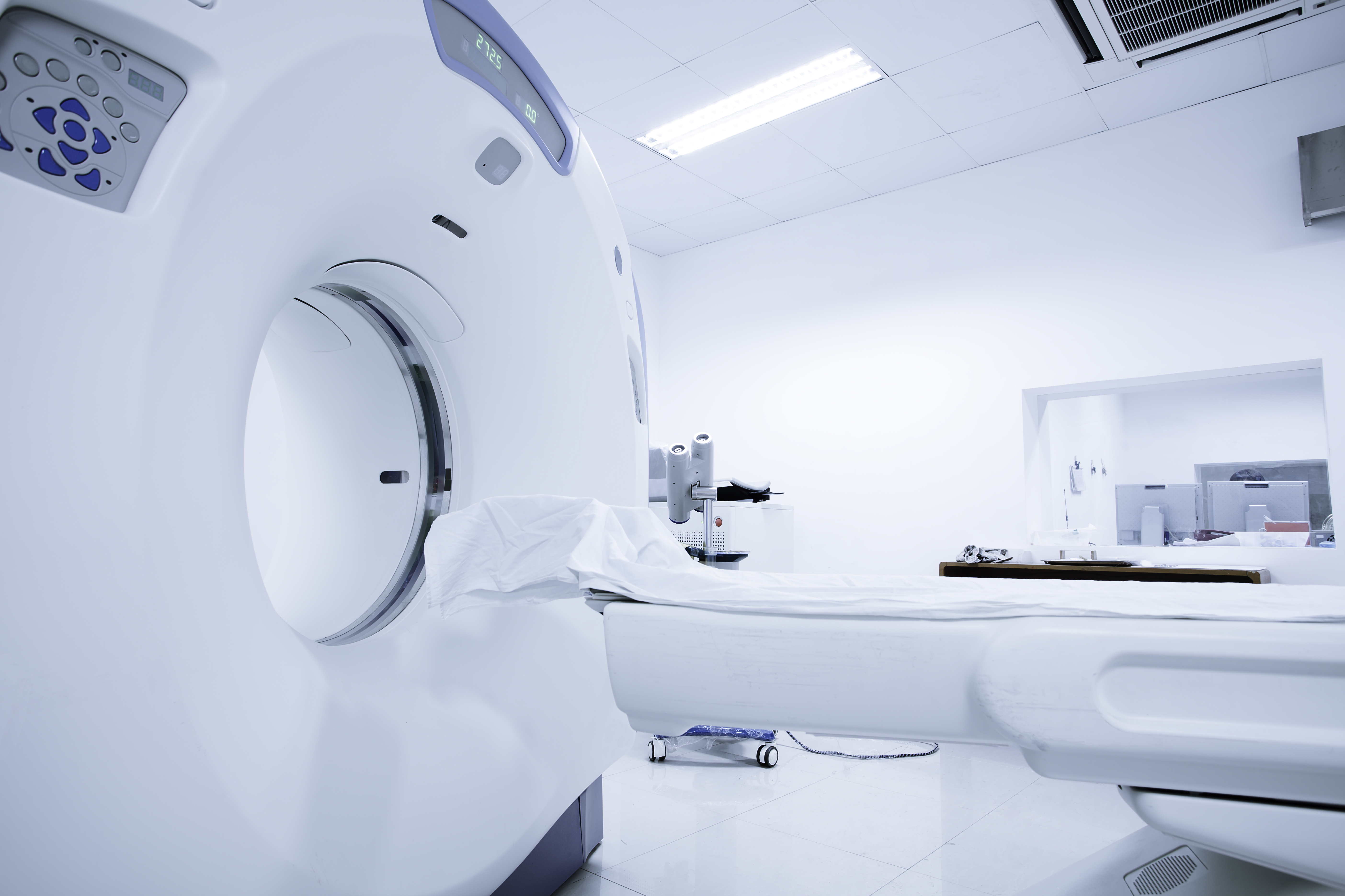 Ein CT-Gerät in einem hellen, modernen Untersuchungraum. Die Maschine hat einen großen, runden Öffnungsbereich und liegt neben einer Liege, die mit einem weißen Laken bedeckt ist. Der Raum hat eine saubere und sterile Atmosphäre.