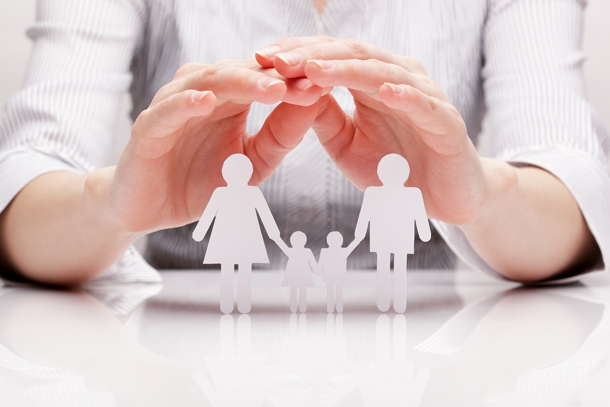 Eine Hand hält schützend über fünf Papierfiguren, die eine Familie darstellen: zwei Erwachsene und zwei Kinder, die sich an den Händen halten. Die Szene vermittelt den Eindruck von Schutz und Zusammenhalt.