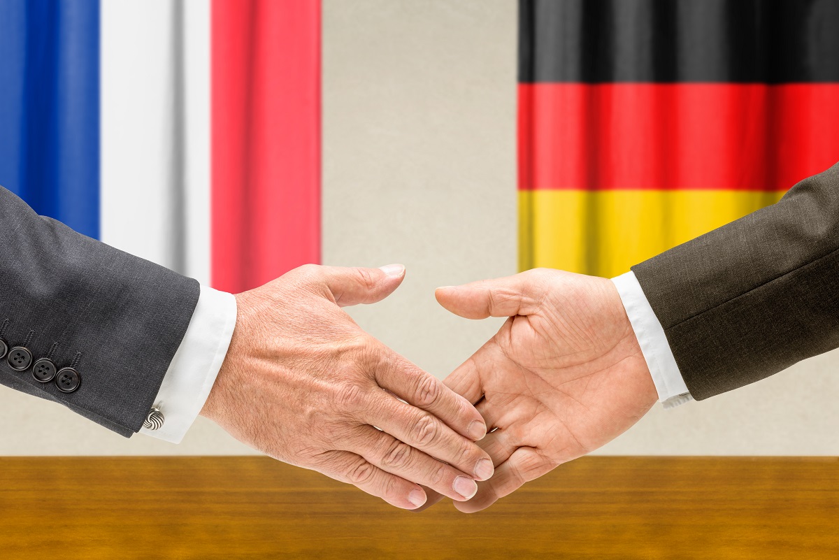 Two hands are shaking over a wooden table, symbolizing agreement or partnership. In the background, blurred flags suggest international cooperation, specifically representing Germany and France. The focus is on the handshake, conveying trust and collaboration.