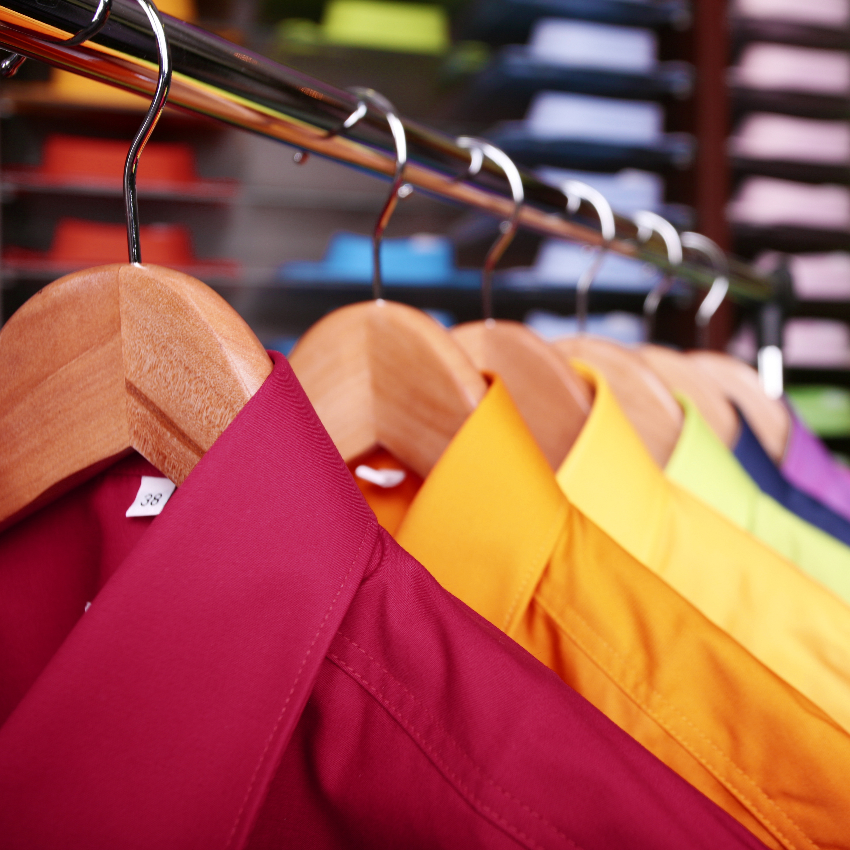 A close-up view of several dress shirts hanging on wooden hangers. The shirts, in shades of burgundy, yellow, and other vibrant colors, showcase a neatly organized rack. The background features blurred stacks of folded clothing.