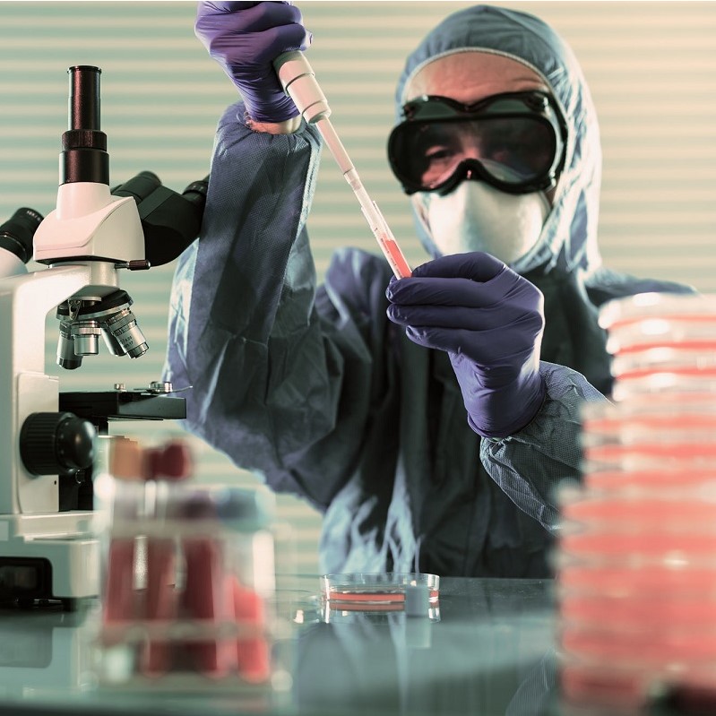 Eine Person in Schutzkleidung hält eine Pipette über ein Reagenzglas und entnimmt eine Flüssigkeit. Im Hintergrund sind ein Mikroskop und mehrere Petrischalen sichtbar, die in unterschiedlichen Farben gefüllt sind. Die Szene spielt sich in einem Labor ab.