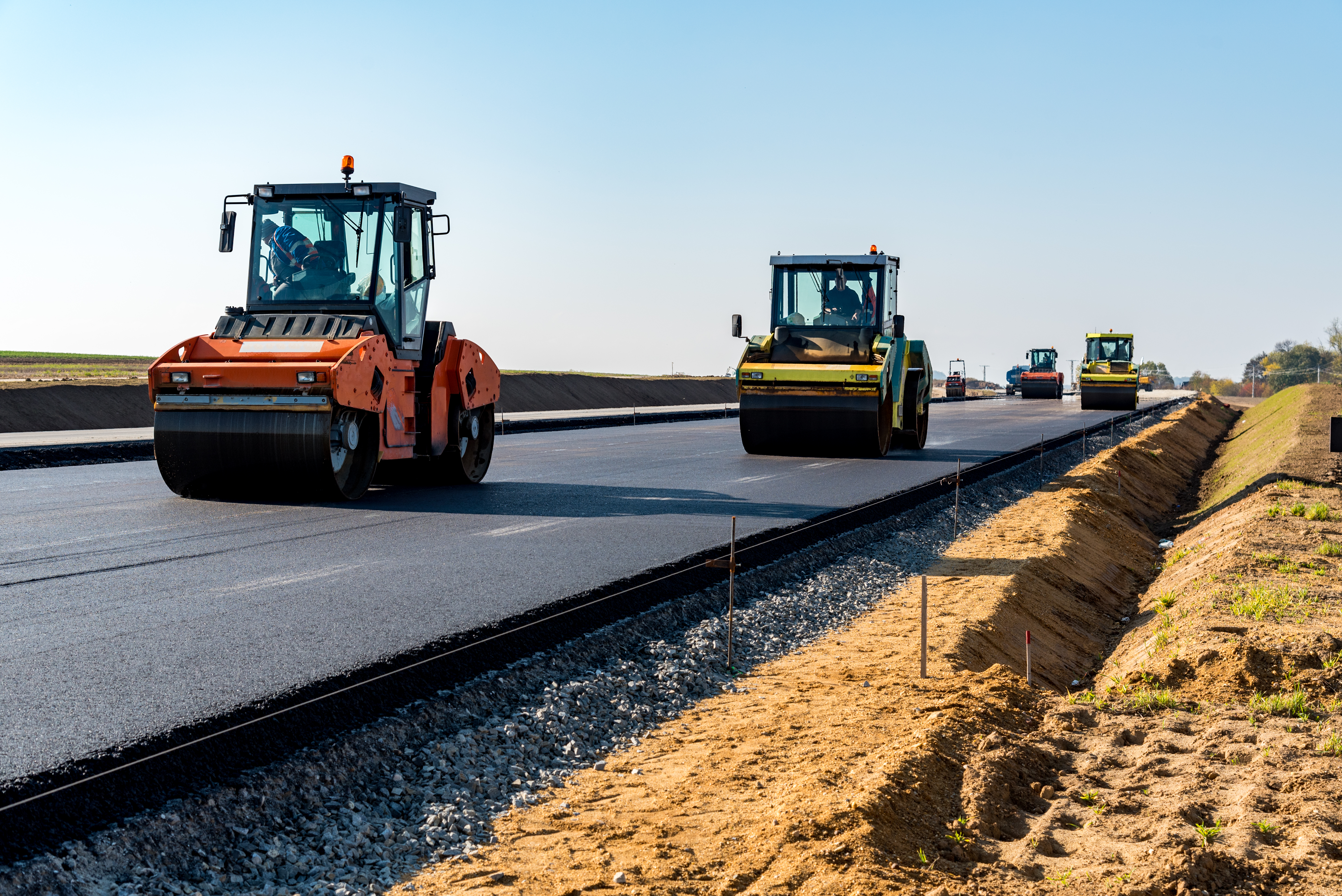 Auf der Straße arbeiten mehrere Straßenwalzen, die frischen Asphalt verdichten. Im Hintergrund sind weitere Baumaschinen zu sehen. Die Szene spielt unter klarem Himmel, was auf einen sonnigen Tag hinweist.
