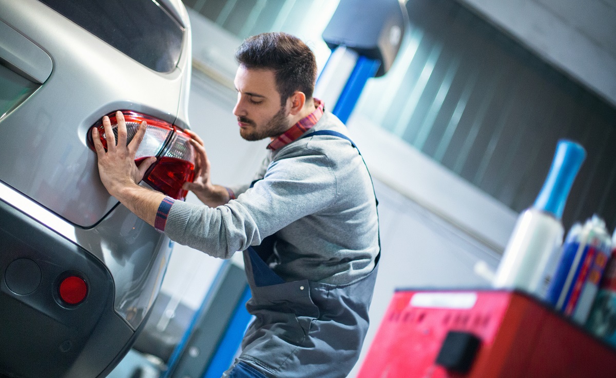 Ein Mechaniker arbeitet an einem Auto und installiert die Rückleuchte. Er konzentriert sich auf seine Aufgabe, während er die Teile des Fahrzeugs prüft. Im Hintergrund sind typische Werkstattutensilien zu sehen.
